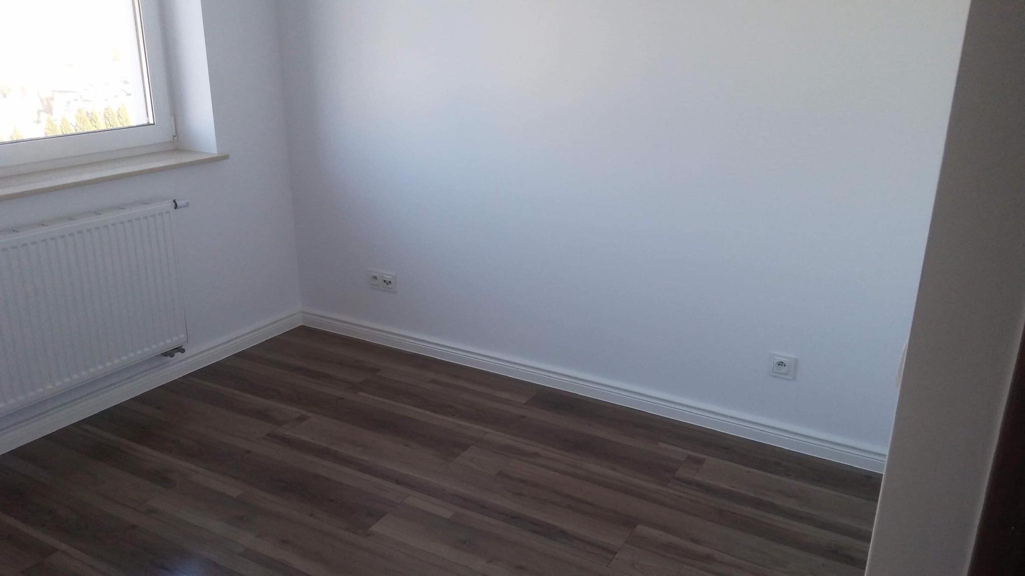 Pomalowane na biało wnętrze pokoju z panelami podłogowymi imitującymi drewno, grzejnikiem pod oknem i gniazdkami elektrycznymi na ścianie.
