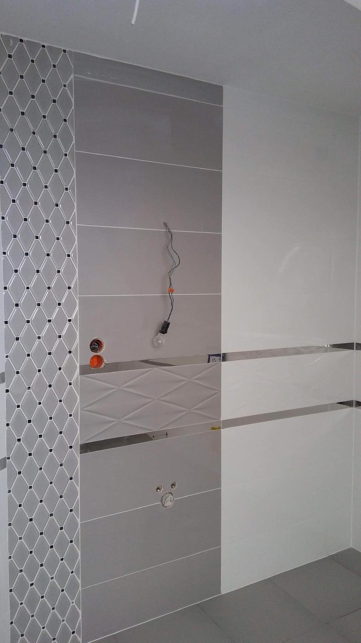 Ściana łazienki w trakcie wykańczania: szare i białe płytki z dekoracyjnym pasem mozaiki, widoczne wystające kable z żarówką i otwory na armaturę.