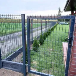 Otwarta furtka z metalowej siatki, część ogrodzenia panelowego, z widokiem na trawnik z tujami i polną drogę.