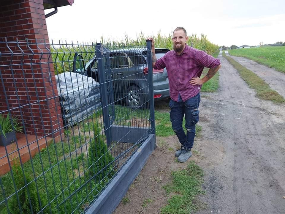 Uśmiechnięty mężczyzna w koszuli w kratę opiera się o nowo zainstalowane, grafitowe ogrodzenie z paneli z podmurówką, z samochodem i zapakowanymi panelami w tle, na wiejskiej drodze.