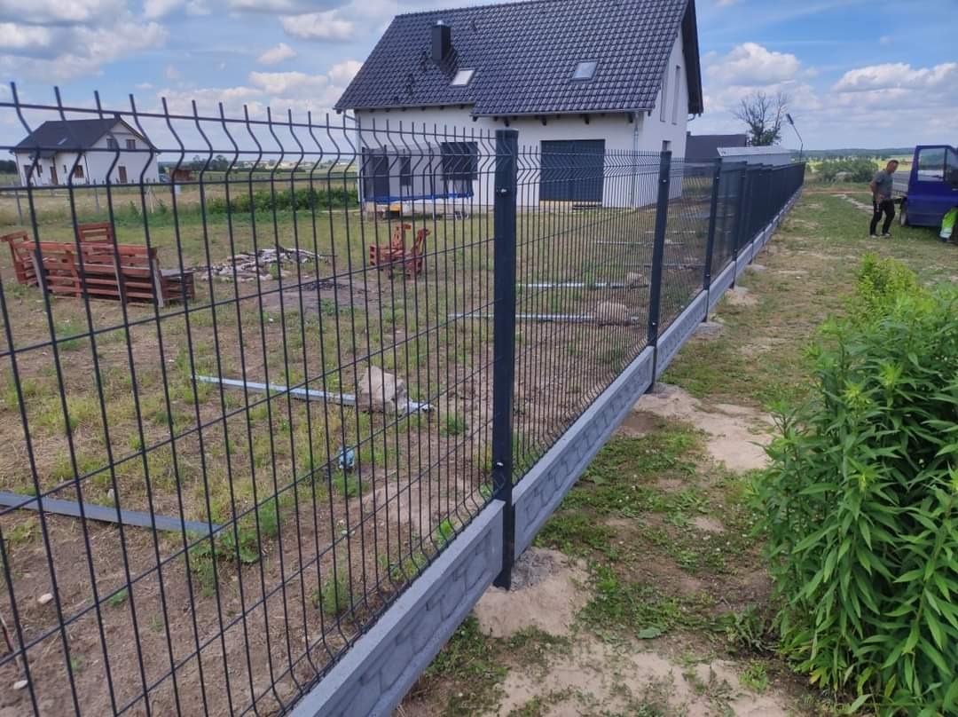 Nowoczesne, czarne ogrodzenie panelowe z podmurówką betonową na tle domu jednorodzinnego i częściowo widocznego samochodu dostawczego.