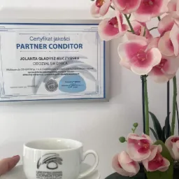 Certyfikat jakości Partner Conditor Jolanty Gładysz-Kuczyńskiej, oddział Świdnica, z filiżanką z logo firmy i dekoracyjną orchideą w tle.
