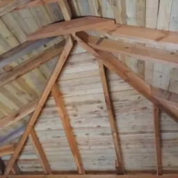 Wnętrze drewnianej konstrukcji dachu z widocznymi elementami więźby i deskowaniem, surowe drewno o naturalnym odcieniu.