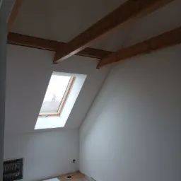 Poddasze z oknem dachowym, widoczne drewniane belki konstrukcyjne pod sufitem, panele podłogowe i instalacja grzewcza w ścianie, oraz fragment listwy przypodłogowej w folii ochronnej.