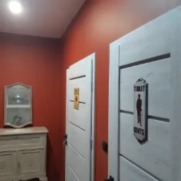 Pomarańczowa ściana z dwoma białymi drzwiami z drewnianą fakturą, na jednych drzwiach znak 'TOILET GENTS' z sylwetką mężczyzny, obok komoda z lustrem.