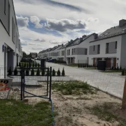 Ulica z rzędem nowych, pomalowanych na biało domów jednorodzinnych z szarymi dachami, widok przez tymczasowe ogrodzenie, z boku widoczny baner reklamowy.