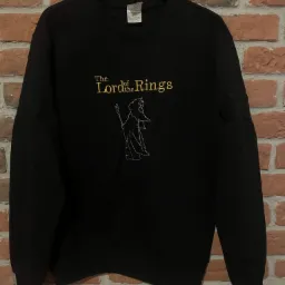 Czarna bluza z haftowanym złotym napisem 'The Lord of the Rings' i konturem postaci czarodzieja, wisząca na wieszaku na tle ceglanej ściany.