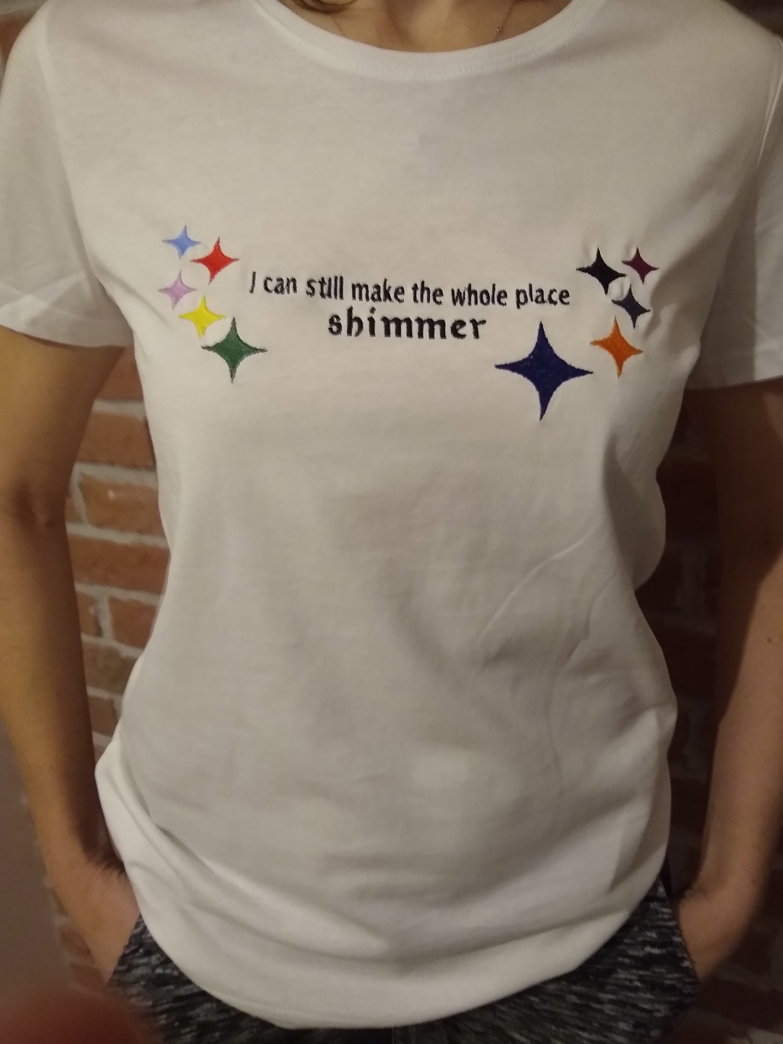 Biała koszulka z krótkim rękawem, ozdobiona kolorowymi, haftowanymi gwiazdami i czarnym napisem 'I can still make the whole place shimmer'.
