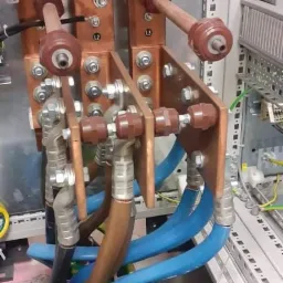 Szczegółowe ujęcie rozdzielnicy elektrycznej z miedzianymi szynami, zabezpieczeniami i podłączonymi przewodami w kolorach niebieskim, czarnym i brązowym.