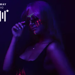 Portret kobiety w okularach, z makijażem neonowym w odcieniach zieleni i czerwieni, oświetlonej fioletowym światłem, na tle logo studia.