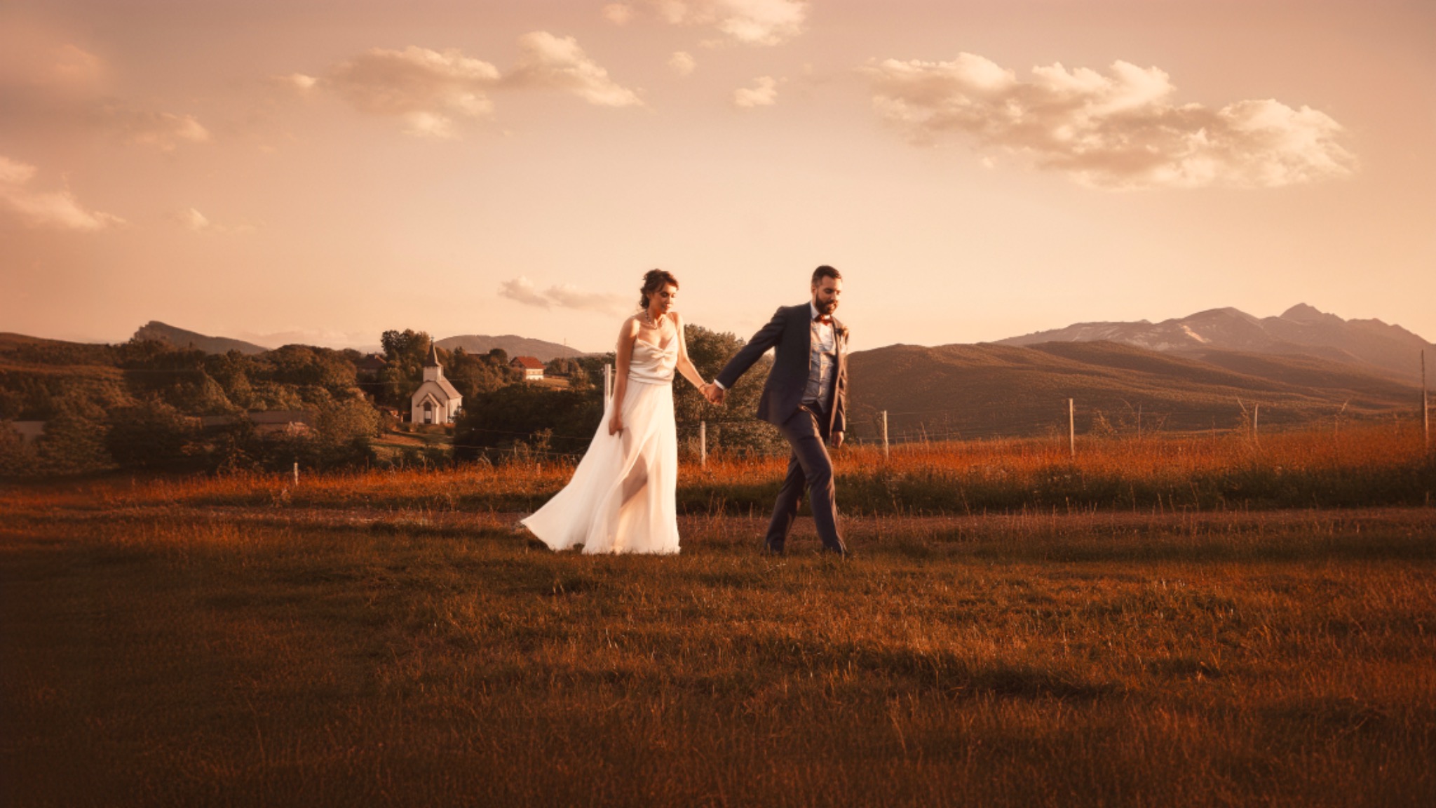 Para młoda trzymająca się za ręce spaceruje po łące w promieniach zachodzącego słońca, w tle kościół i góry.