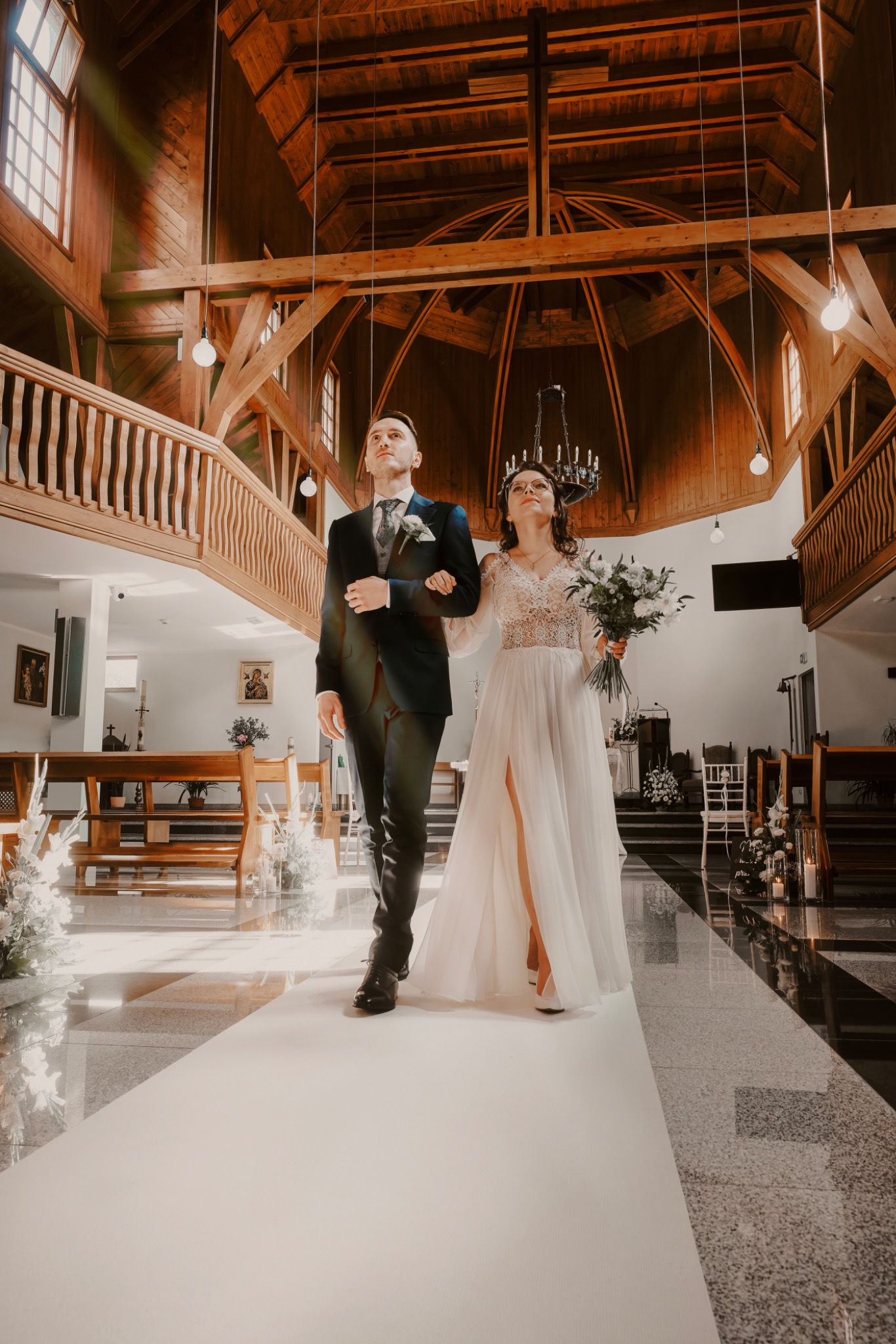 Para młoda, panna młoda w sukni z koronkową górą i rozcięciem, pan młody w ciemnym garniturze, idą po białym dywanie w kościele z drewnianą architekturą.