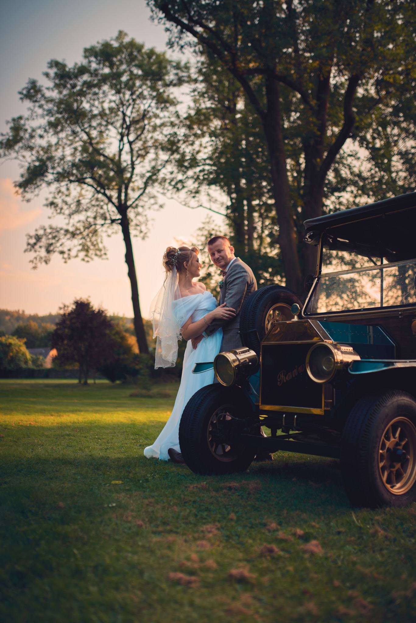 Para młoda w objęciach pozuje przy zabytkowym, ciemnozielonym samochodzie na tle zielonego trawnika i drzew w promieniach zachodzącego słońca.