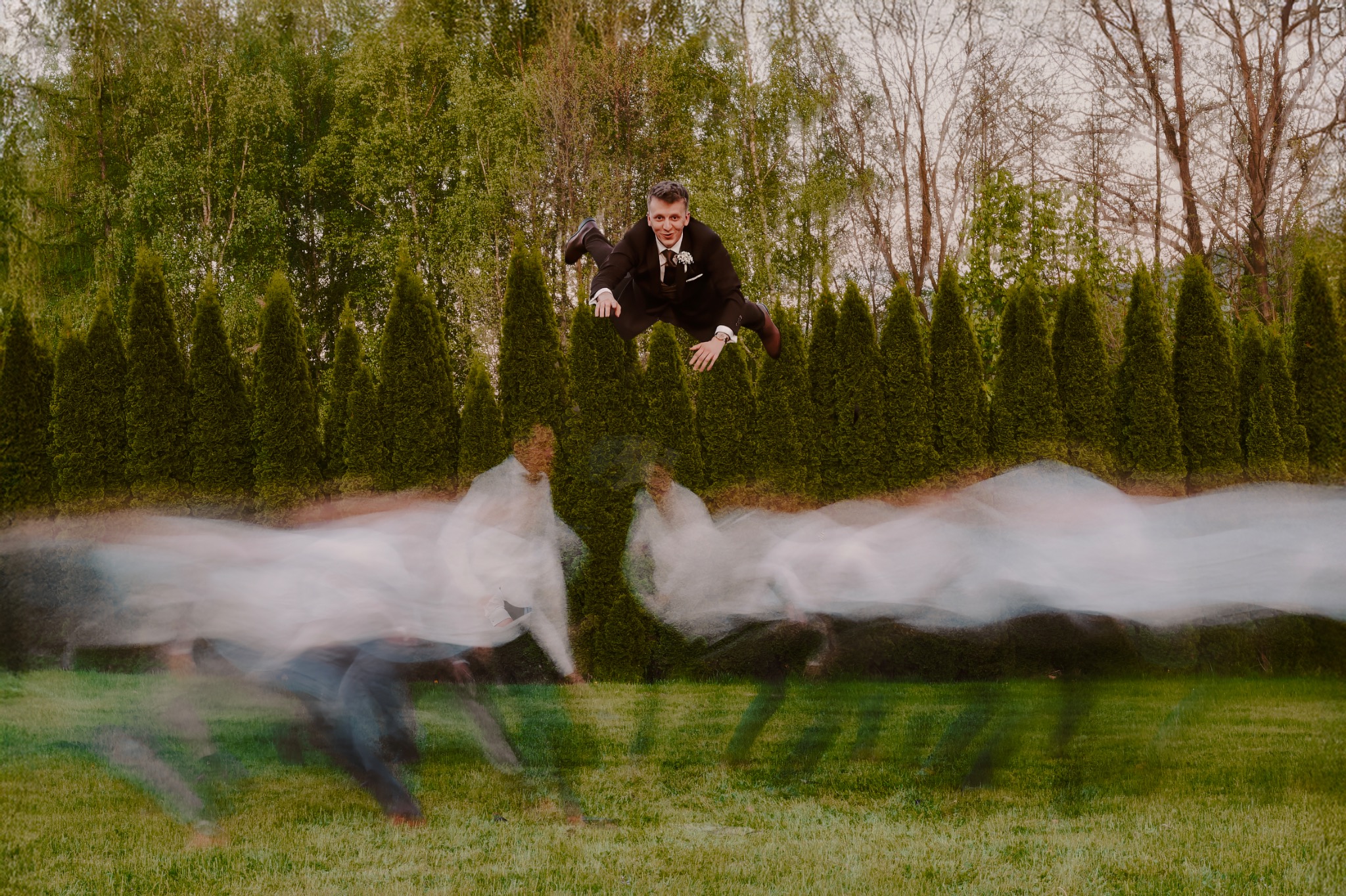 Dynamiczne ujęcie pana młodego w locie na tle zieleni, z efektem rozmycia ruchu i drzew w tle. Nietypowa fotografia ślubna w plenerze.