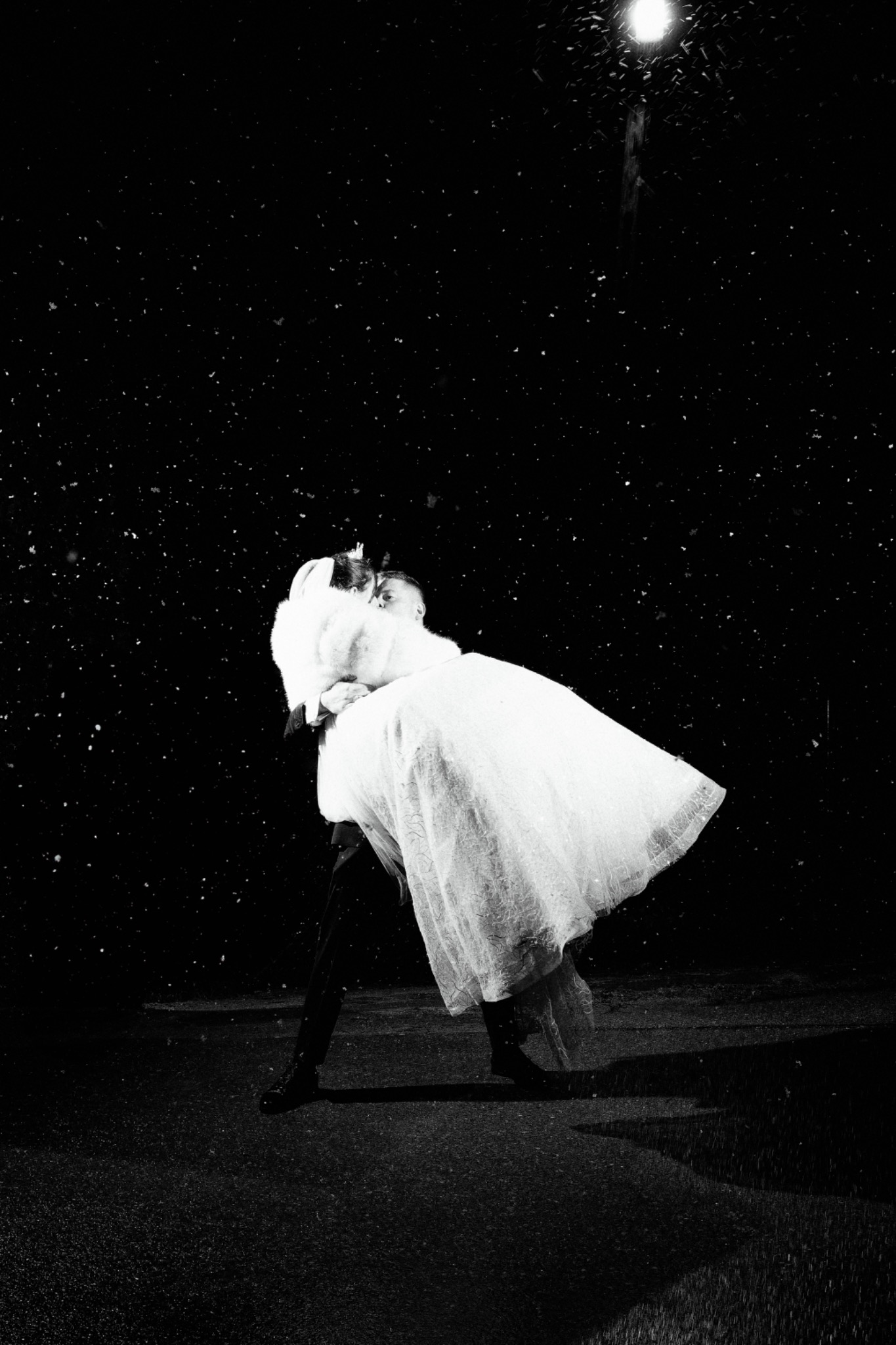 Czarno-białe, artystyczne zdjęcie pary młodej w tańcu pod latarnią podczas opadów. Panna młoda w zwiewnej sukni i futrze, pan młody w ciemnym garniturze. Nastrojowe oświetlenie.