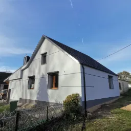 Dom jednorodzinny po remoncie elewacji, widoczna świeżo pomalowana fasada w jasnoszarym odcieniu z ciemnoszarą podmurówką, czarne okna z drewnianymi ramami, błękitne niebo w tle.