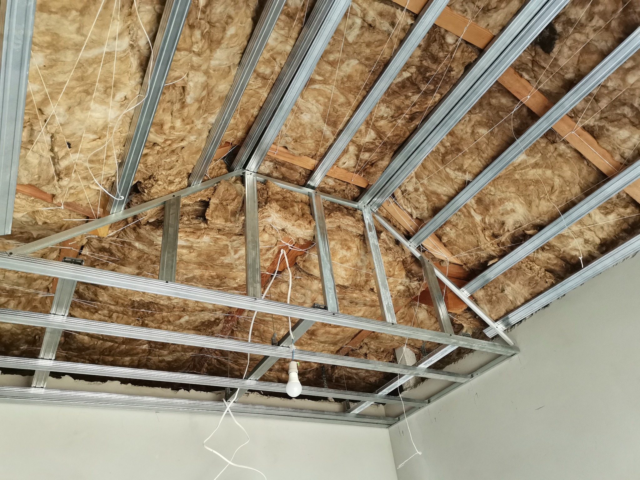 Skośny sufit poddasza w trakcie izolacji wełną mineralną, widoczna metalowa konstrukcja nośna i prowizoryczne oświetlenie żarówką.