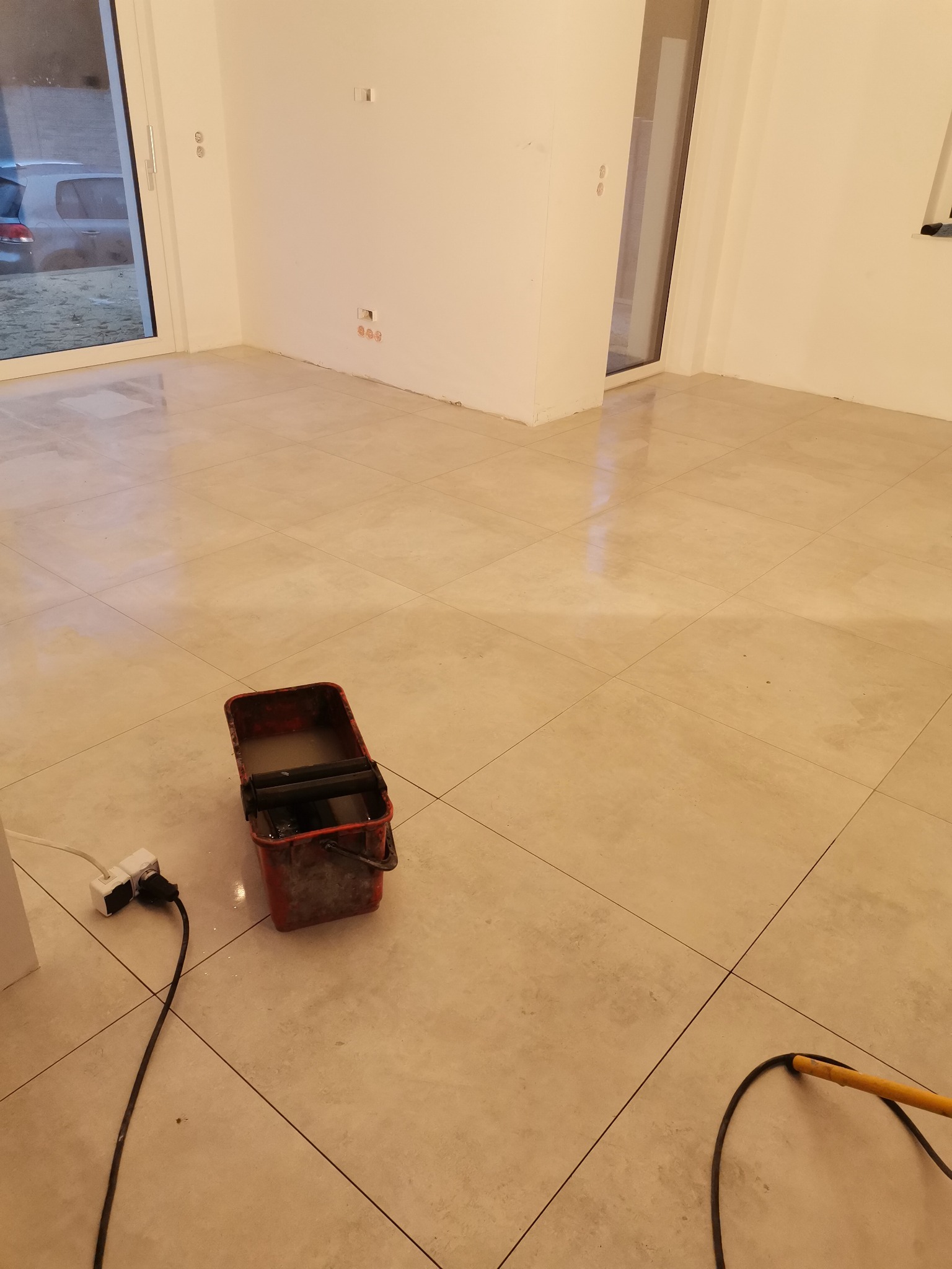 Świeżo położona, jasna, kwadratowa podłoga z płytek ceramicznych, wiadro z wodą i mop obok ściany z widocznymi gniazdkami elektrycznymi i otworami po kontakcie.