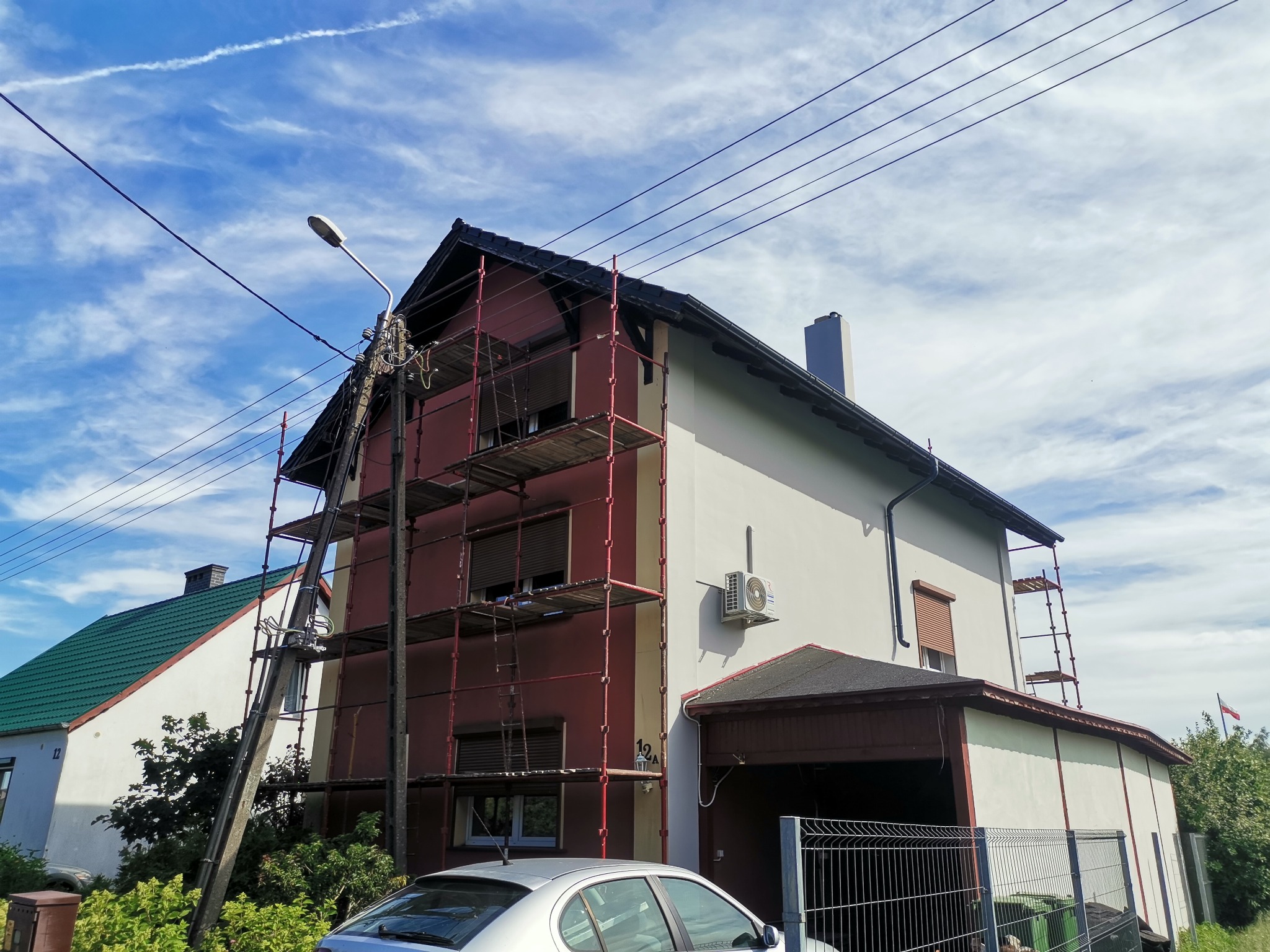 Dom jednorodzinny w trakcie prac elewacyjnych, widoczne rusztowania na tle błękitnego nieba z smugami kondensacyjnymi.