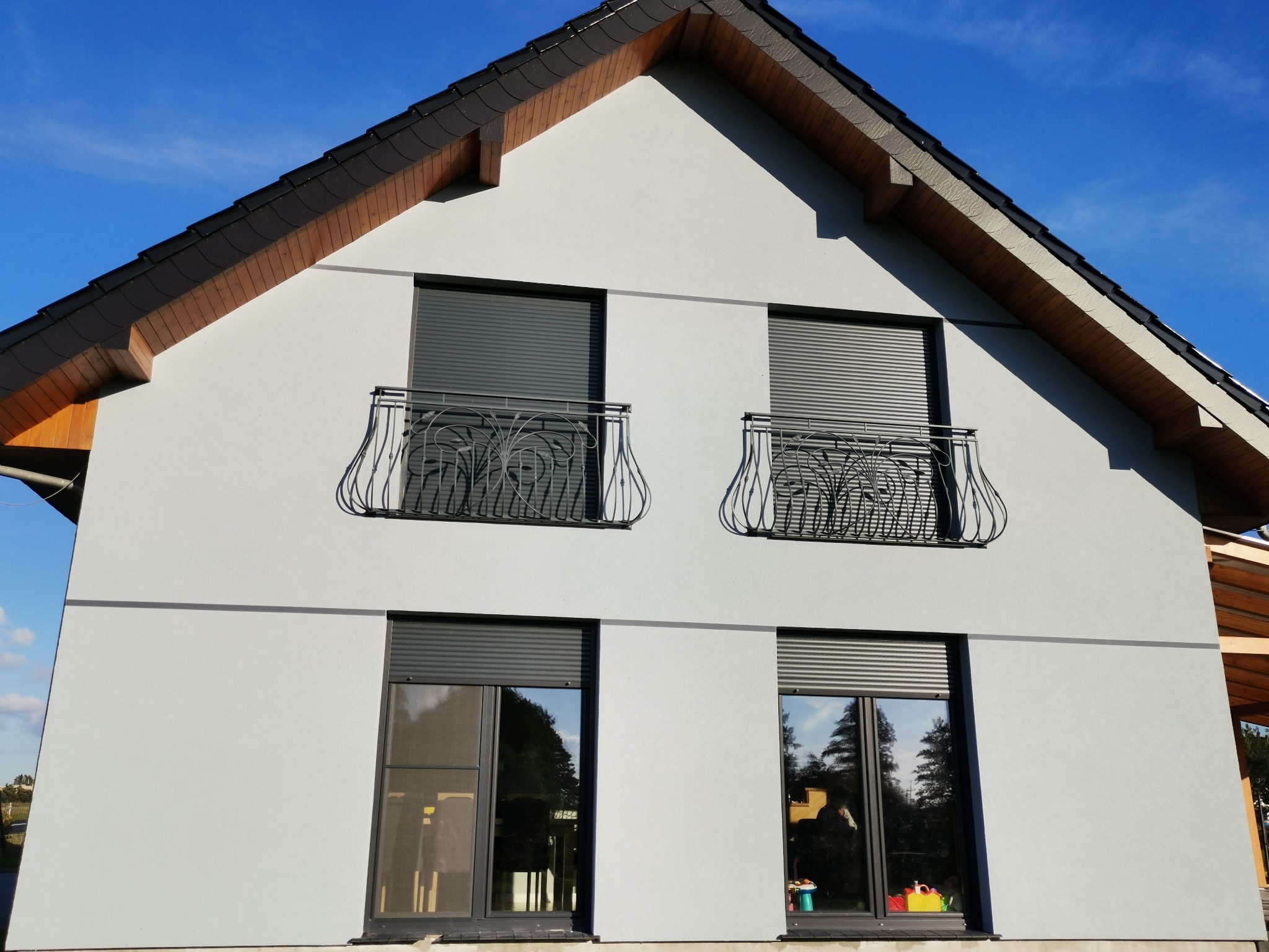 Dwupiętrowy dom z szarą elewacją, czarnymi roletami i ozdobnymi balkonami, sfotografowany w słoneczny dzień.