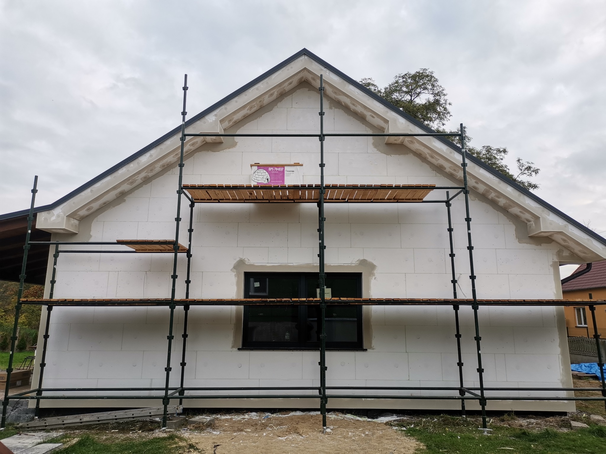 Budynek jednorodzinny w trakcie prac ociepleniowych elewacji, widoczne rusztowanie, białe płyty styropianowe oraz częściowo wykończony fragment szczytu z widocznym oknem.