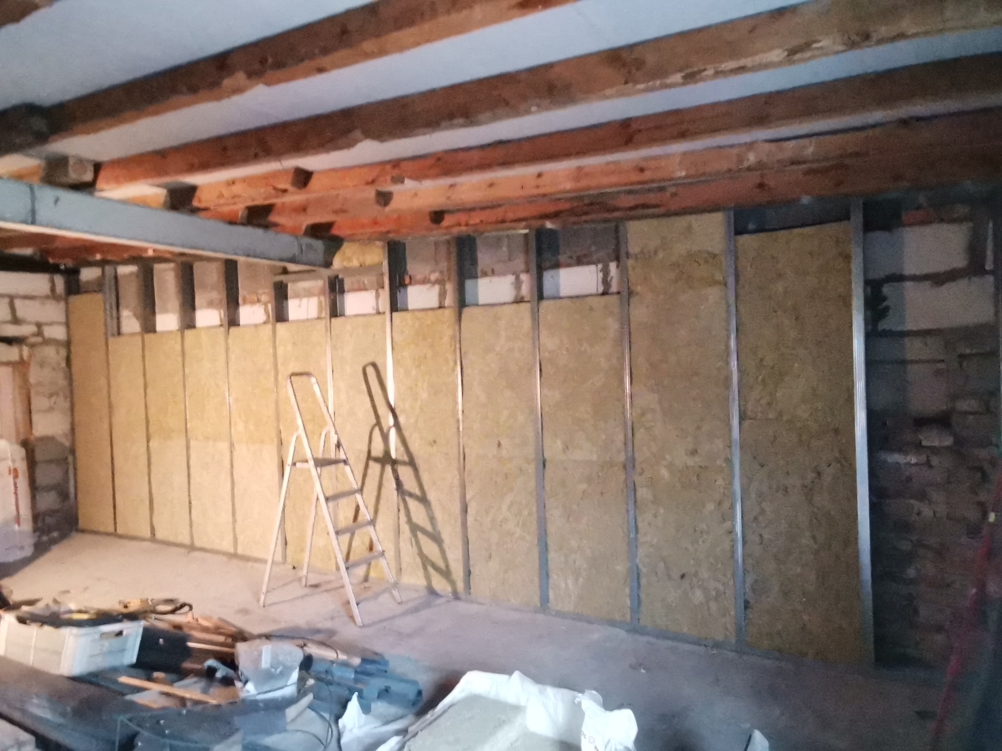 Wewnętrzne ściany z izolacją z wełny mineralnej w metalowej ramie, widoczna drabina i narzędzia w surowym wnętrzu, drewniany strop