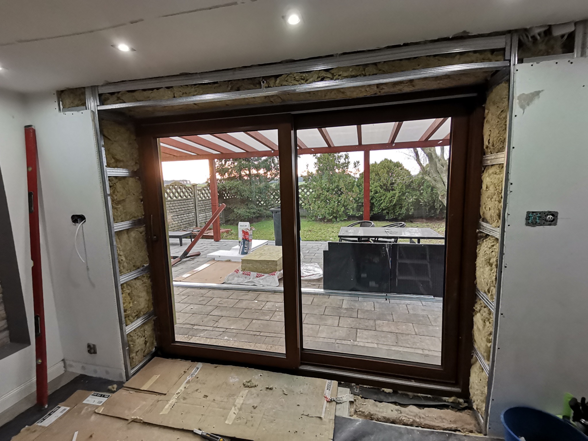 Montaż przesuwnych drzwi tarasowych z widoczną izolacją ścian wełną mineralną i konstrukcją metalową, poziomica przy ścianie, widok na taras z kostki brukowej i ogród.