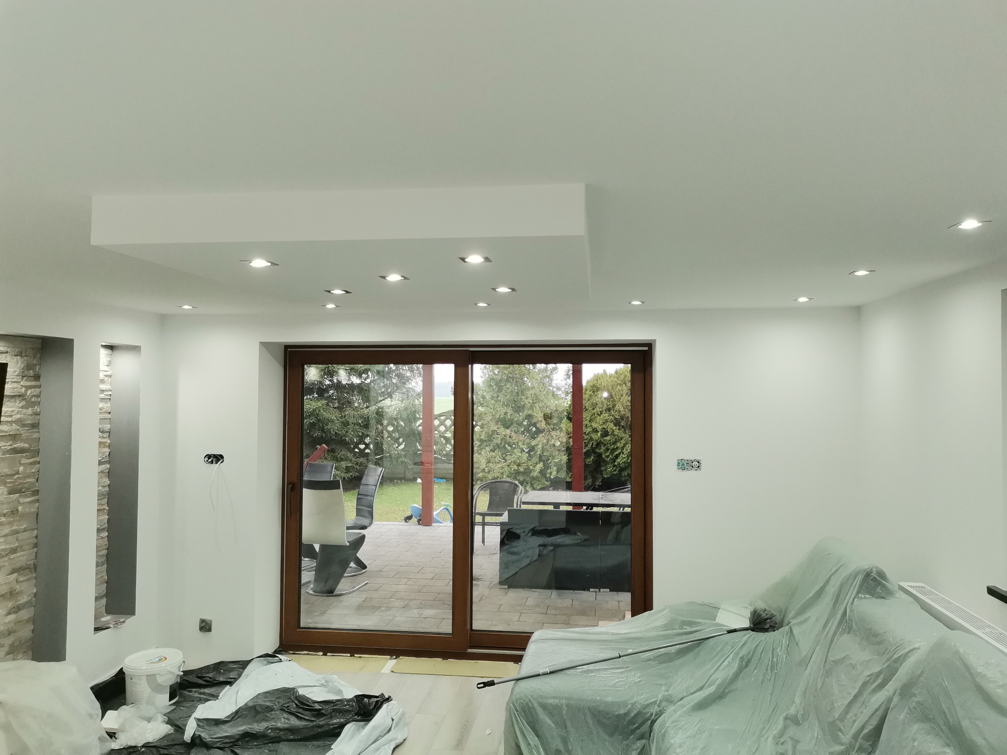 Przygotowanie do malowania ścian w Rostarzewie: meble zabezpieczone folią malarską, widoczne puszki z farbą i odsłonięte gniazdka elektryczne, duże okno przesuwne z widokiem na taras.