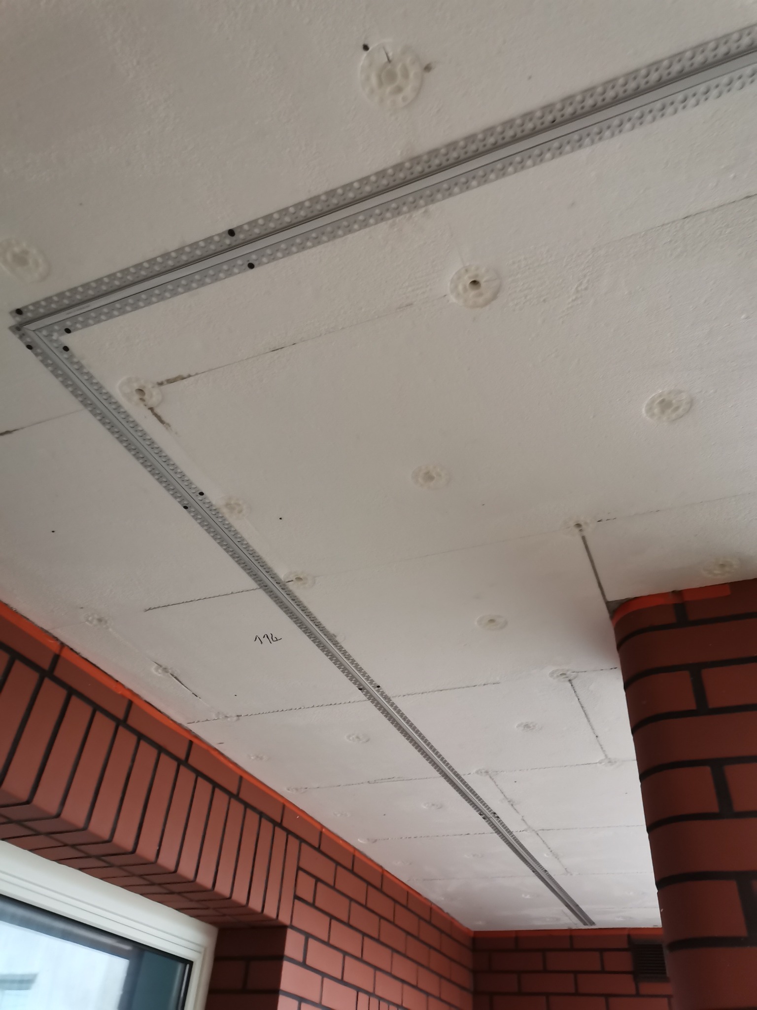 Białe płyty styropianowe zamontowane na suficie z widocznymi metalowymi profilami mocującymi i wypustkami kleju, ceglana ściana w tle.