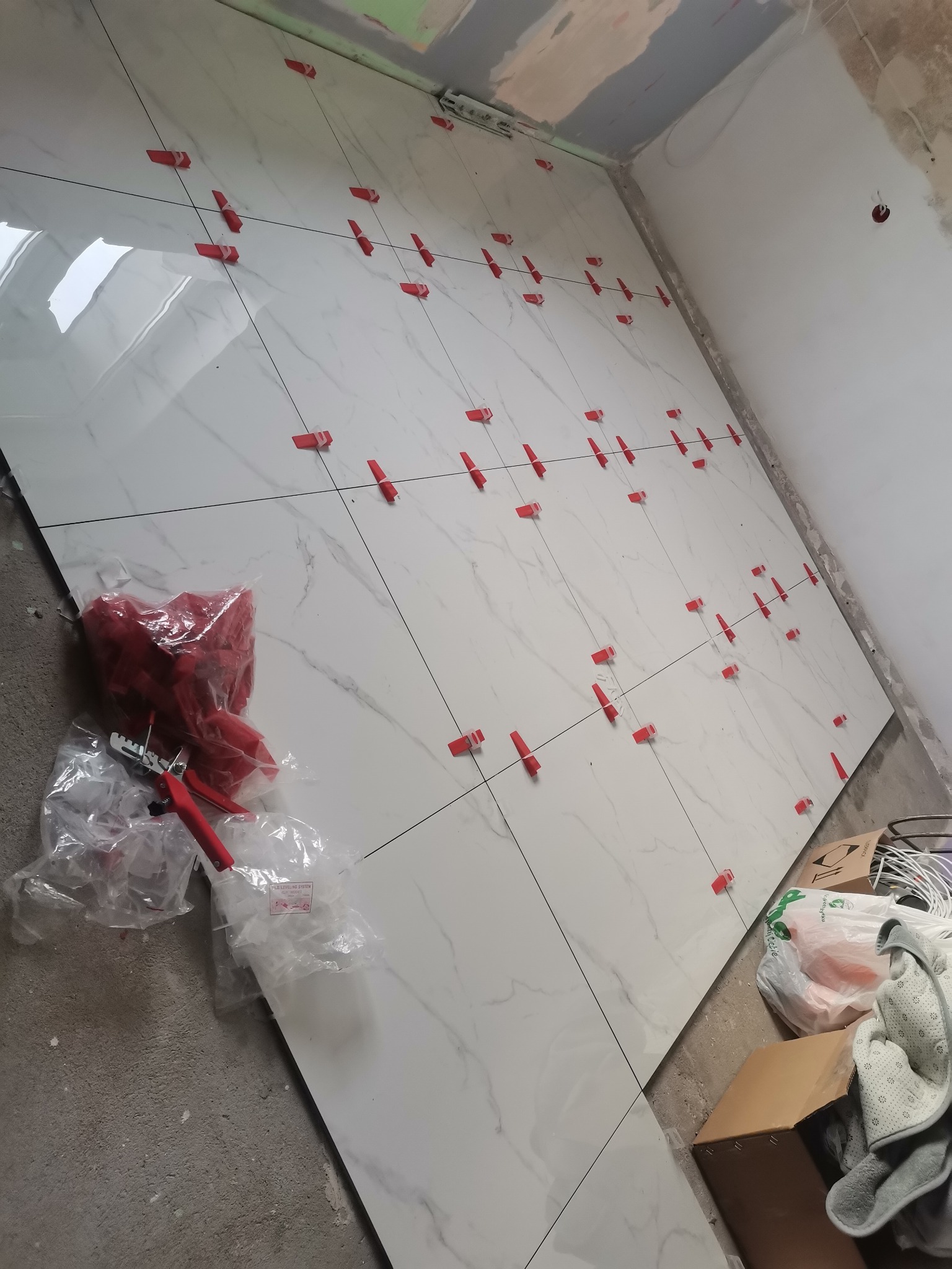 Układanie białych, marmurkowych płytek na podłodze, z użyciem czerwonych klipsów poziomujących i narzędzi w plastikowych opakowaniach, widoczne niedokończone ściany.