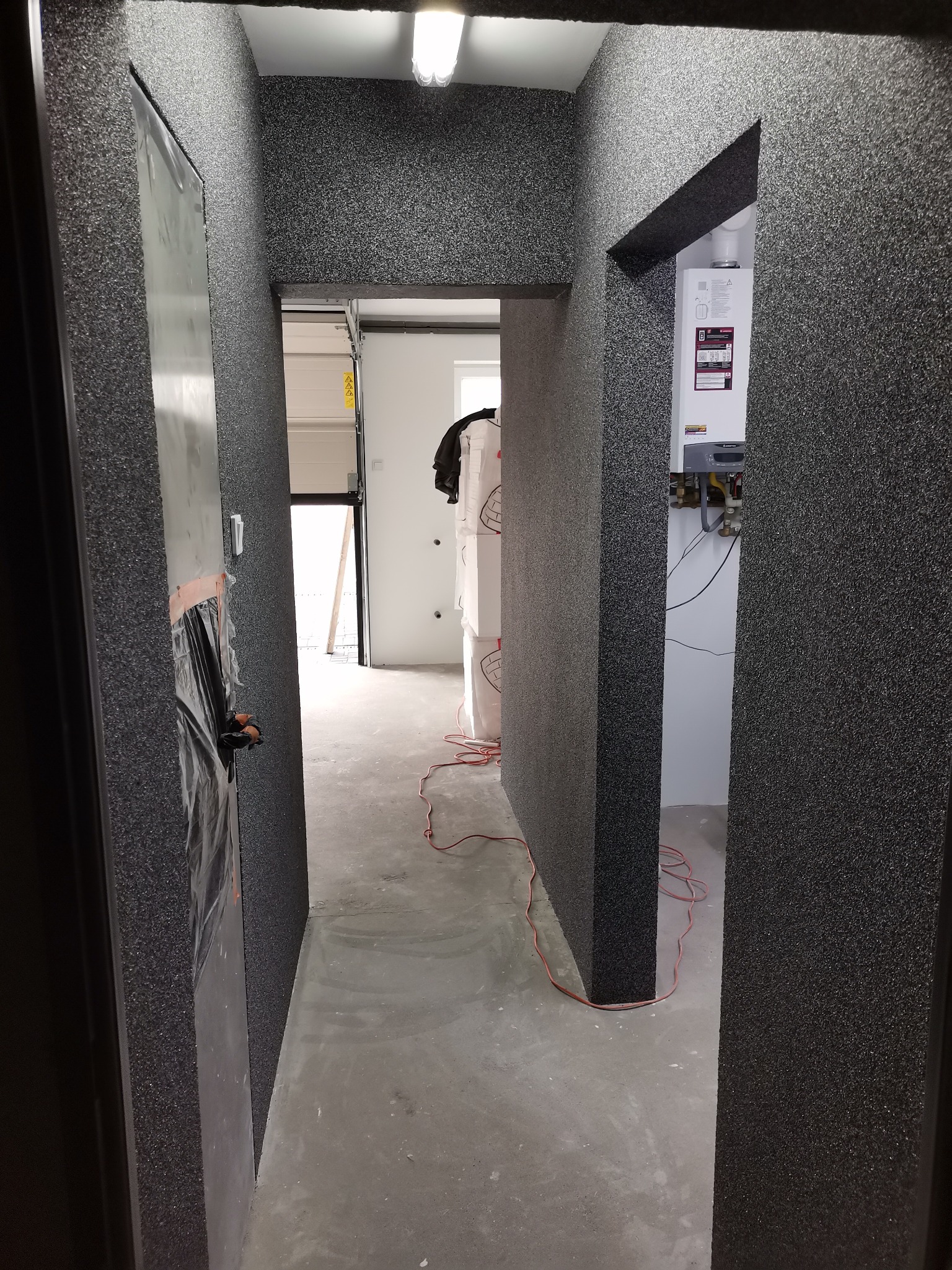 Korytarz z wykończonymi ścianami w ciemnej, ziarnistej fakturze, widoczny bojler na ścianie w głębi, drzwi po lewej stronie częściowo zakryte folią ochronną, podłoga surowa betonowa, widok...