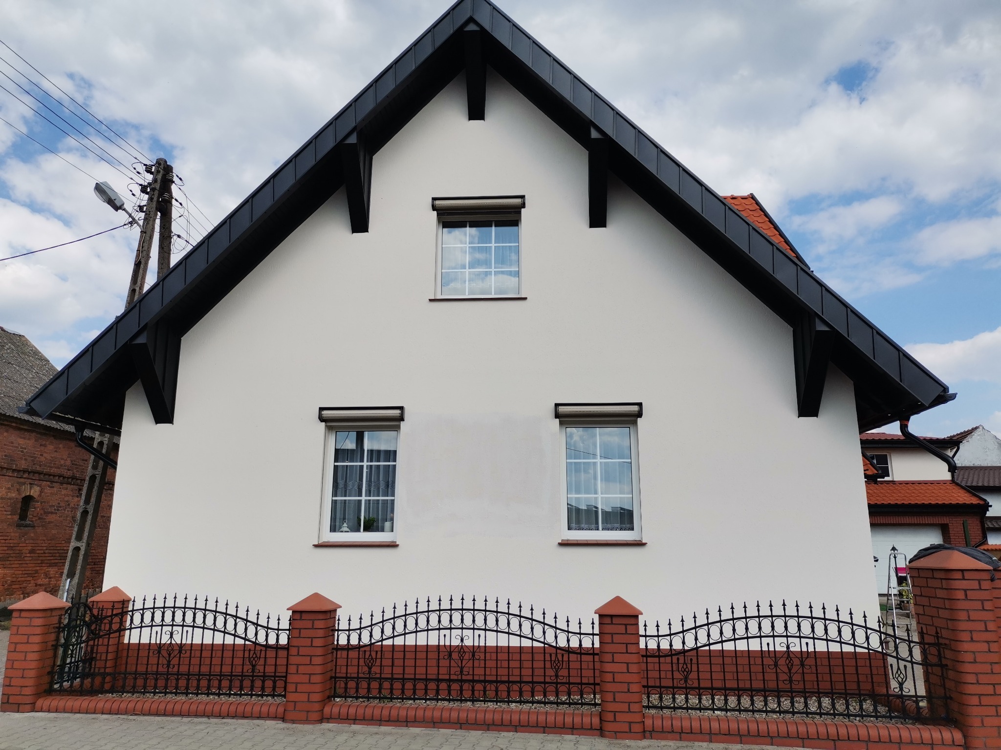 Świeżo wykończona, biała elewacja domu jednorodzinnego z czarnym dachem i trzema oknami z roletami, ogrodzona niskim, kutym płotem z ceglanymi słupkami.