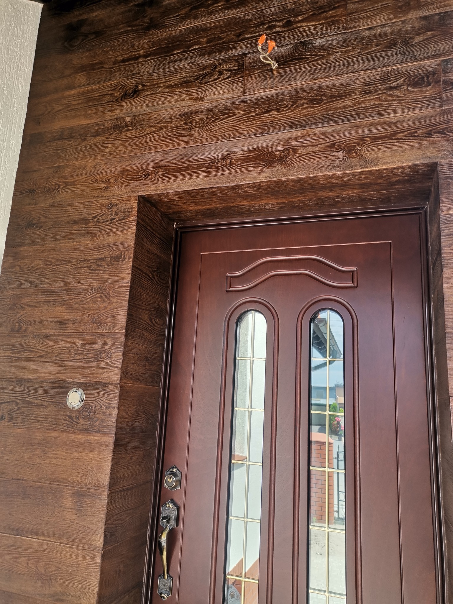 Wejście do domu z brązowymi drzwiami z szybkami i elewacją imitującą drewno. Widoczne wystające kable elektryczne.