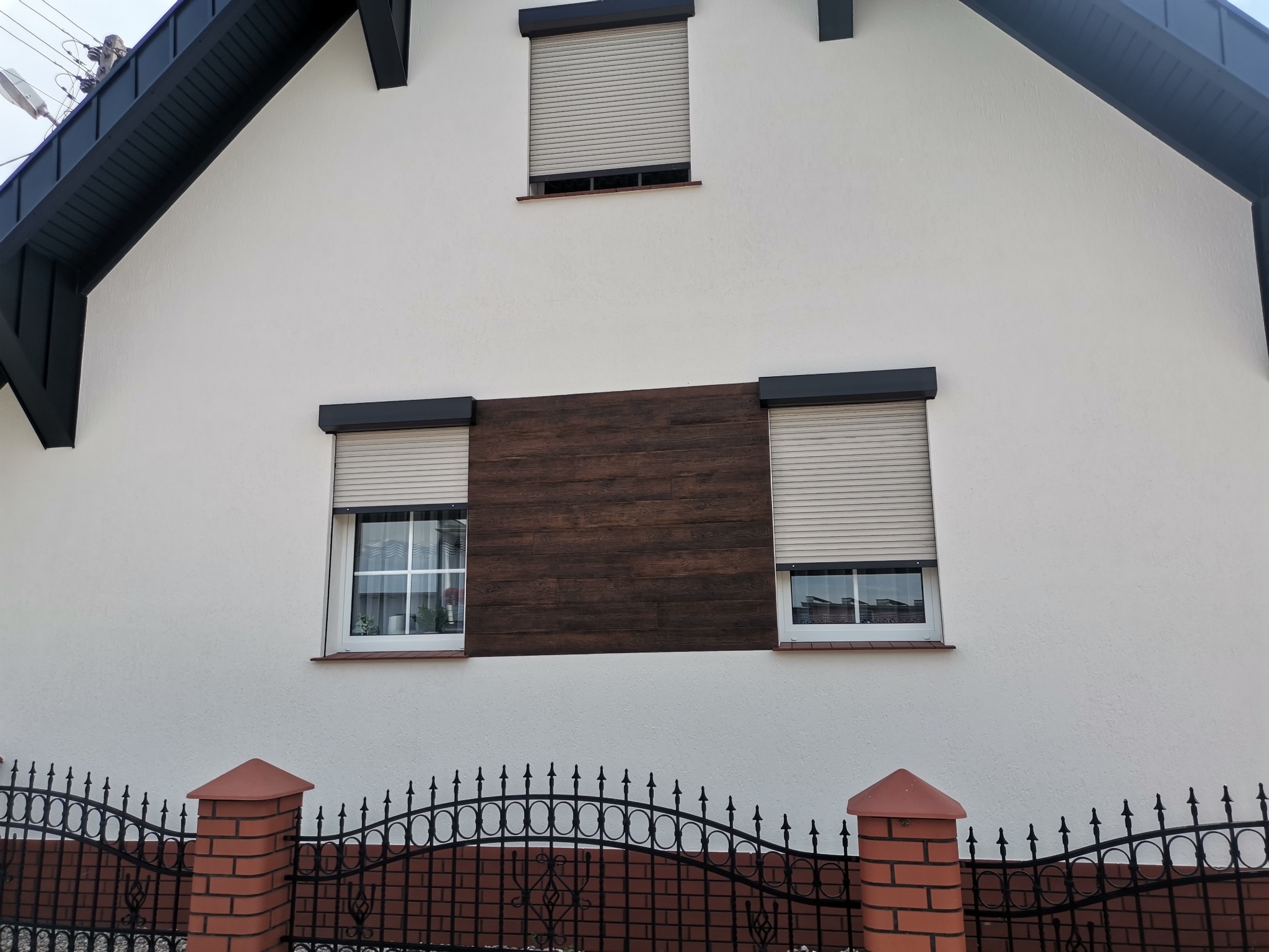 Biała elewacja domu z drewnianym panelem między oknami i roletami zewnętrznymi, widoczny fragment ogrodzenia z ceglanymi słupkami.