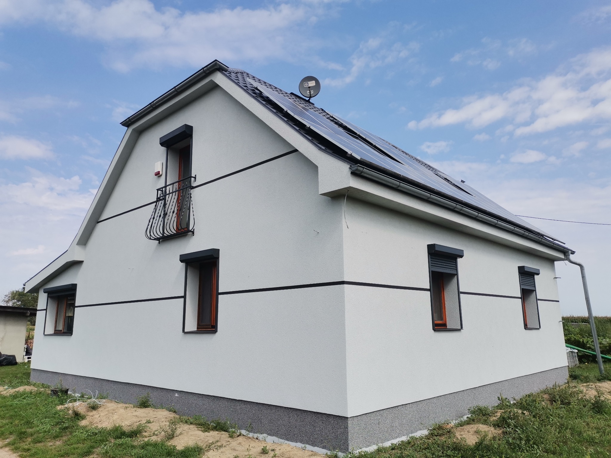 Nowoczesny dom jednorodzinny w Rostarzewie z jasną elewacją, ciemnymi okiennicami, balkonem z kutą balustradą i panelami słonecznymi na dachu.