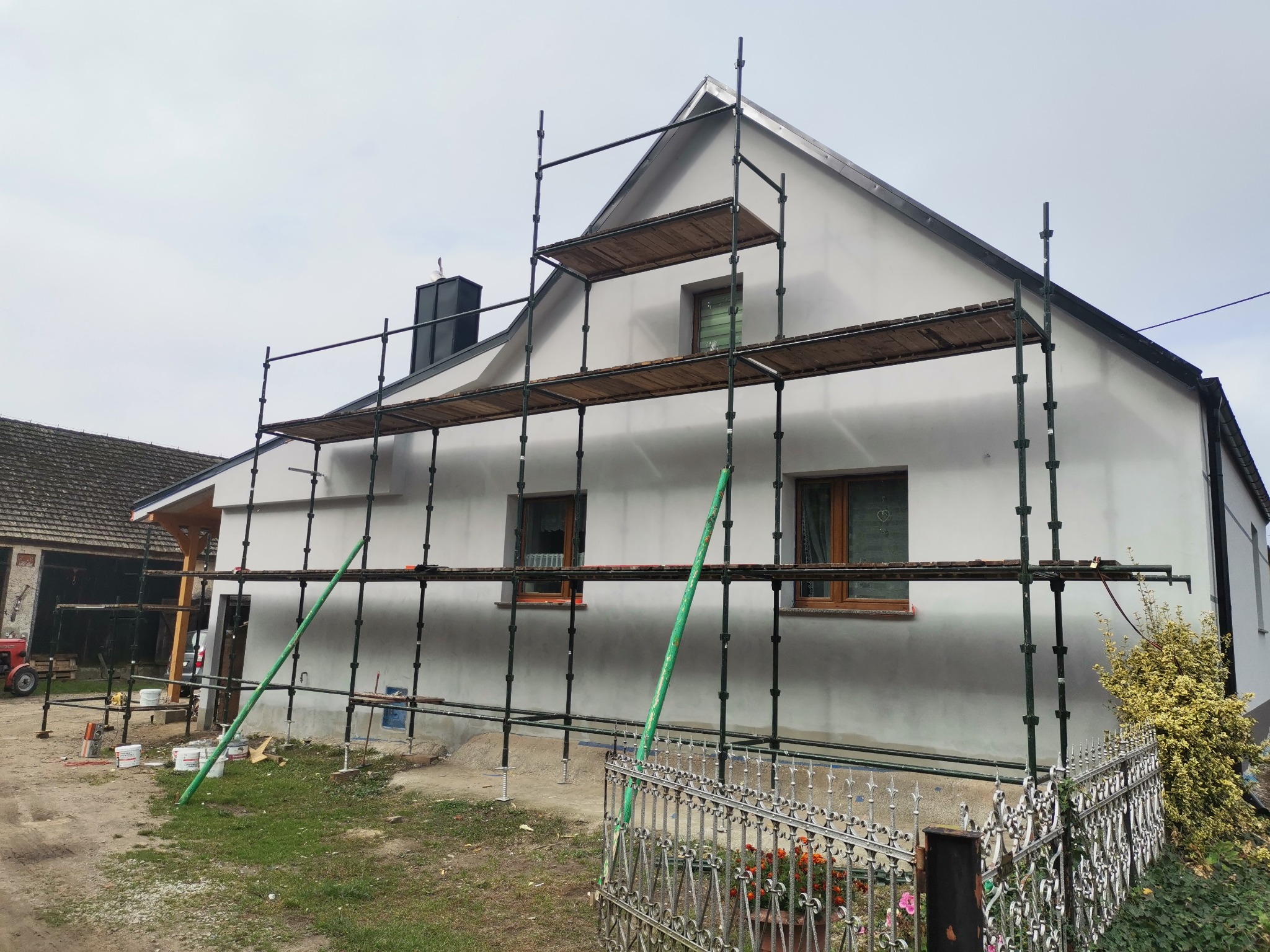 Dom jednorodzinny w trakcie ocieplania elewacji, widoczne rusztowania z drewnianymi pomostami, ściany pokryte warstwą ociepleniową, na ziemi wiadra z zaprawą.