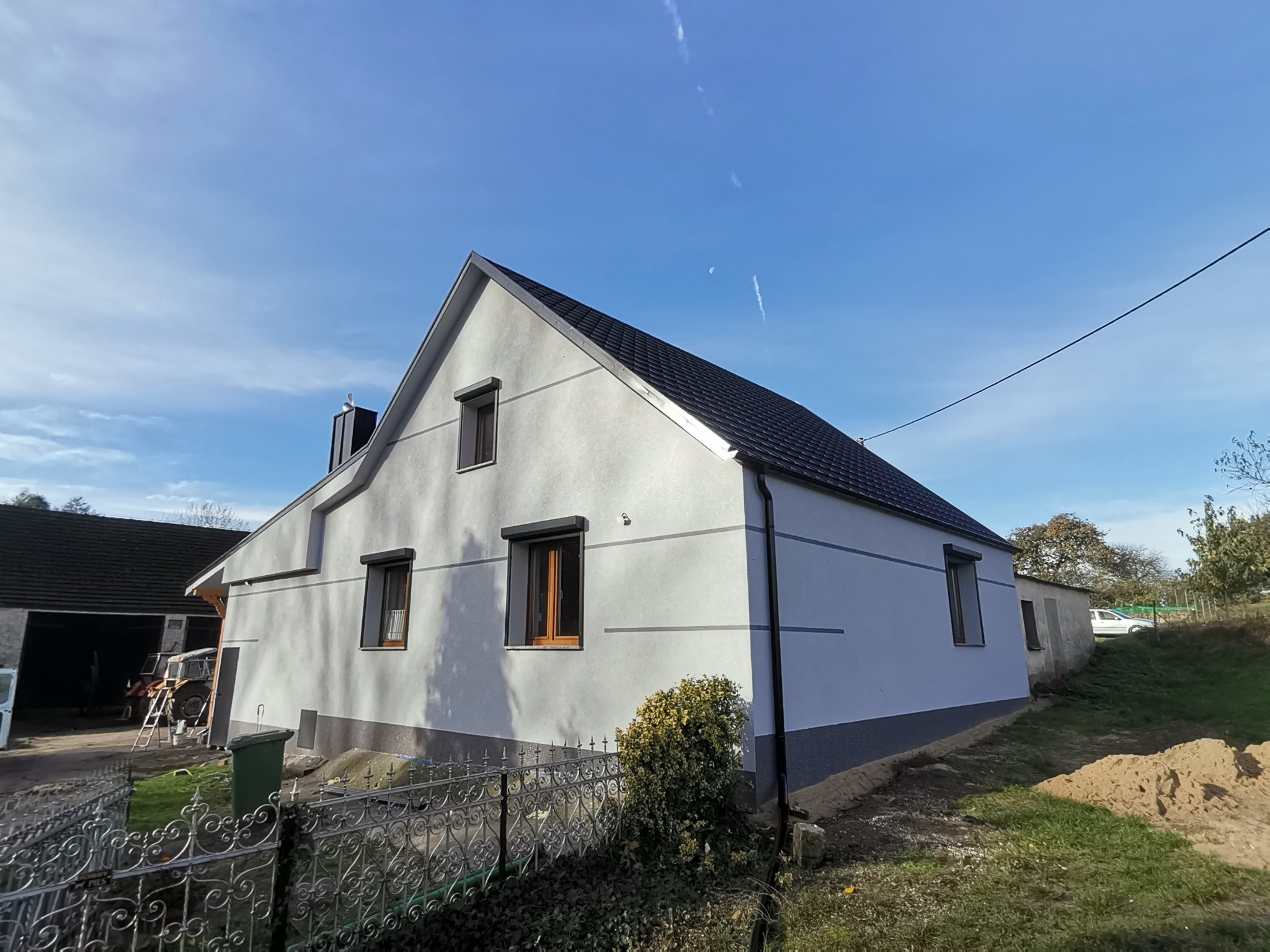 Dom jednorodzinny po remoncie elewacji, widoczna świeżo pomalowana fasada w jasnoszarym odcieniu z ciemnoszarą podmurówką, czarne okna z drewnianymi ramami, błękitne niebo w tle.