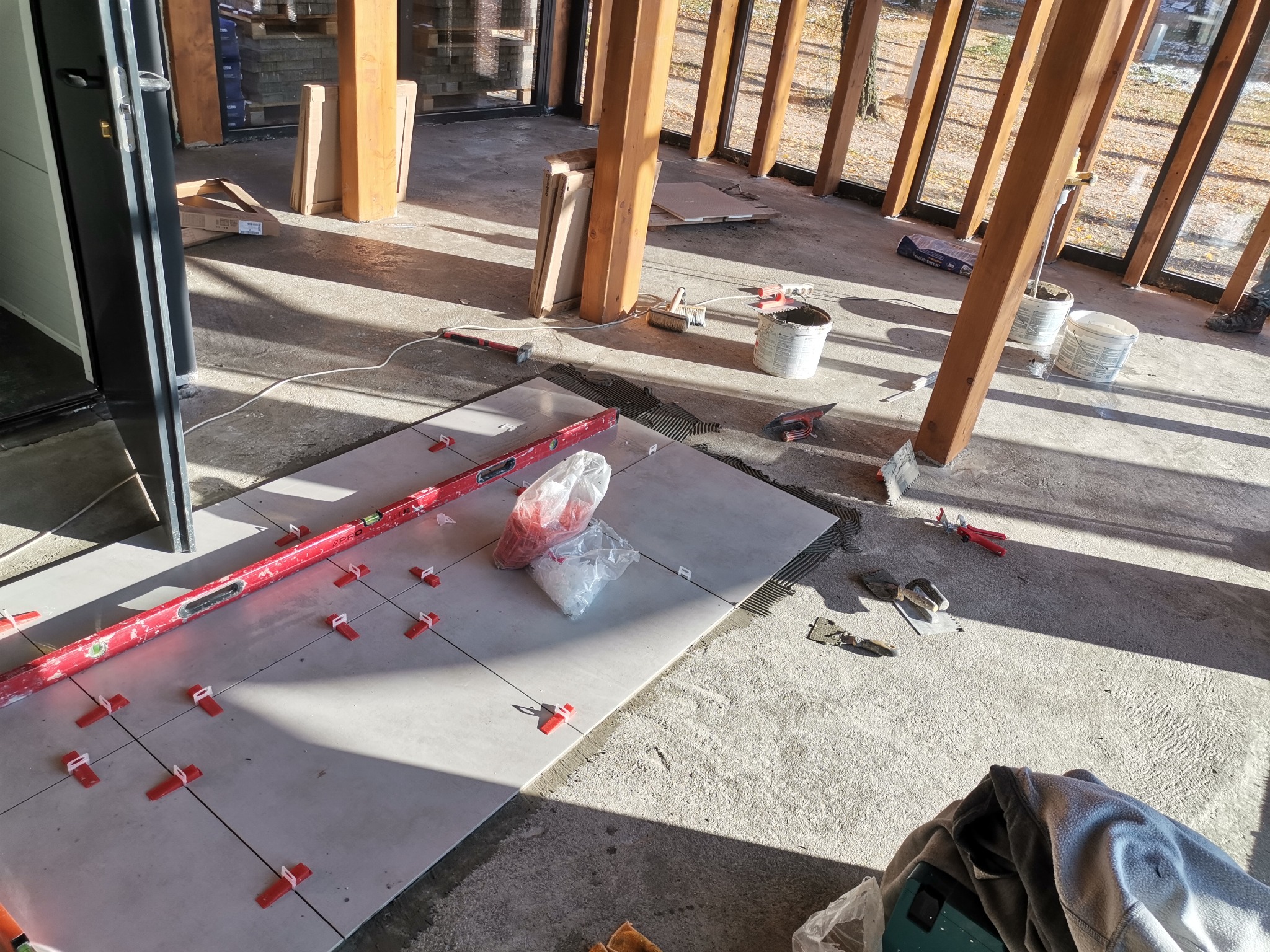 Układanie jasnoszarych płytek podłogowych w przeszklonym pomieszczeniu, widoczna poziomica, krzyżyki dystansowe, woreczki z zaprawą i narzędzia glazurnicze.