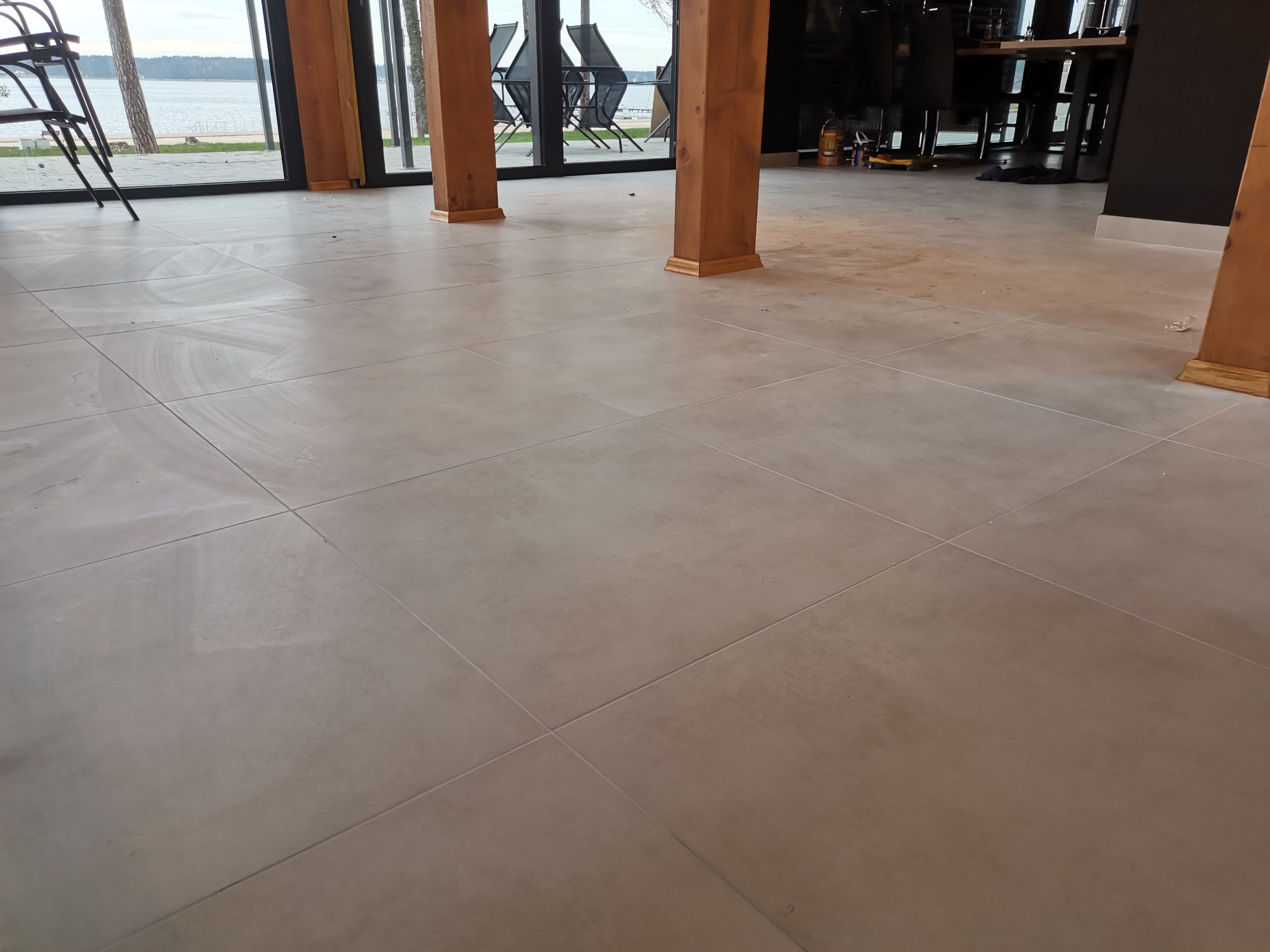 Jasnoszara podłoga z płytek w przestronnym wnętrzu z drewnianymi słupami i widokiem na jezioro za oknami.