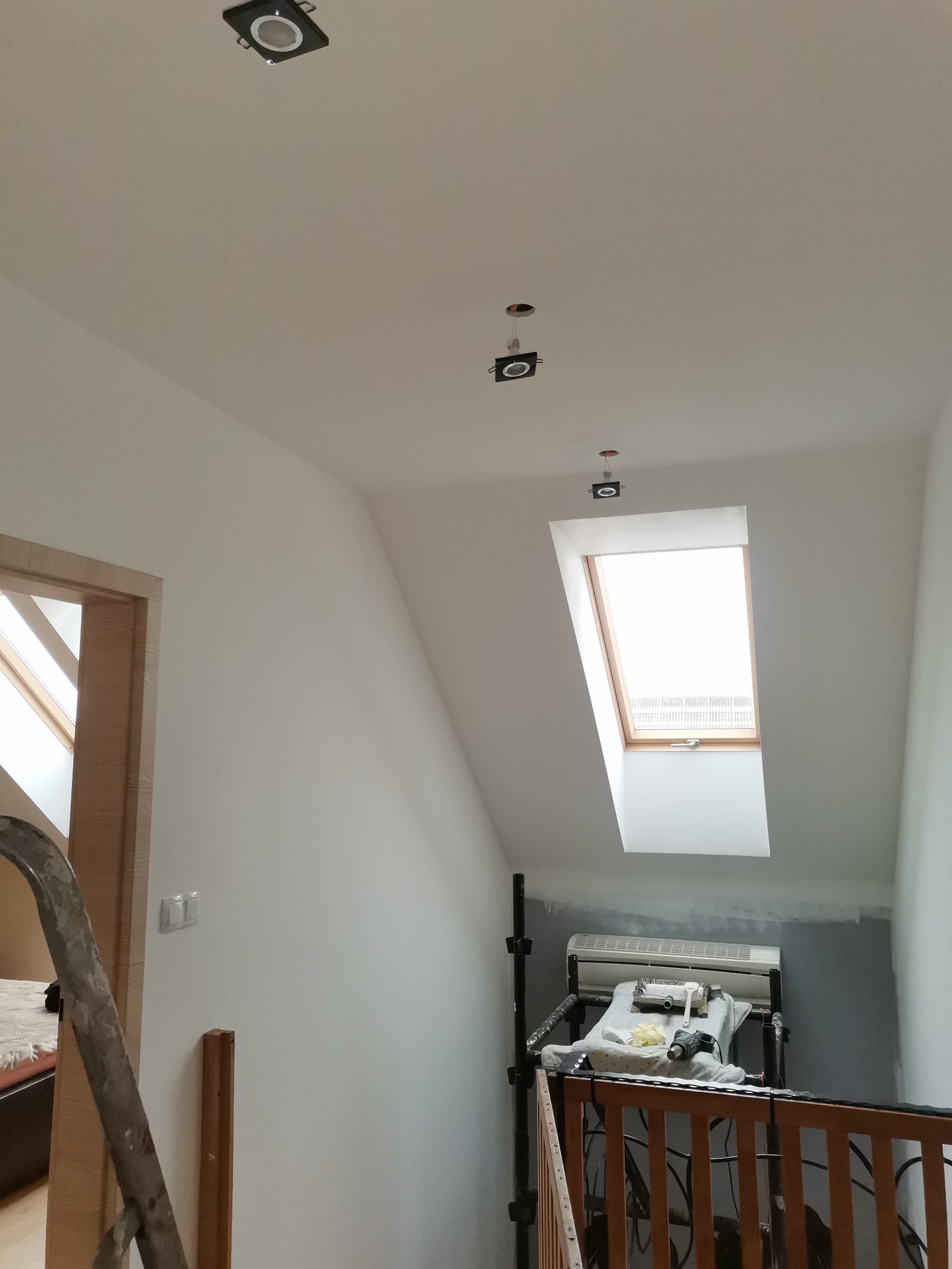 Poddasze po malowaniu na biało, widoczne okno dachowe, zamontowane oprawy oświetleniowe i drabina malarska z narzędziami.