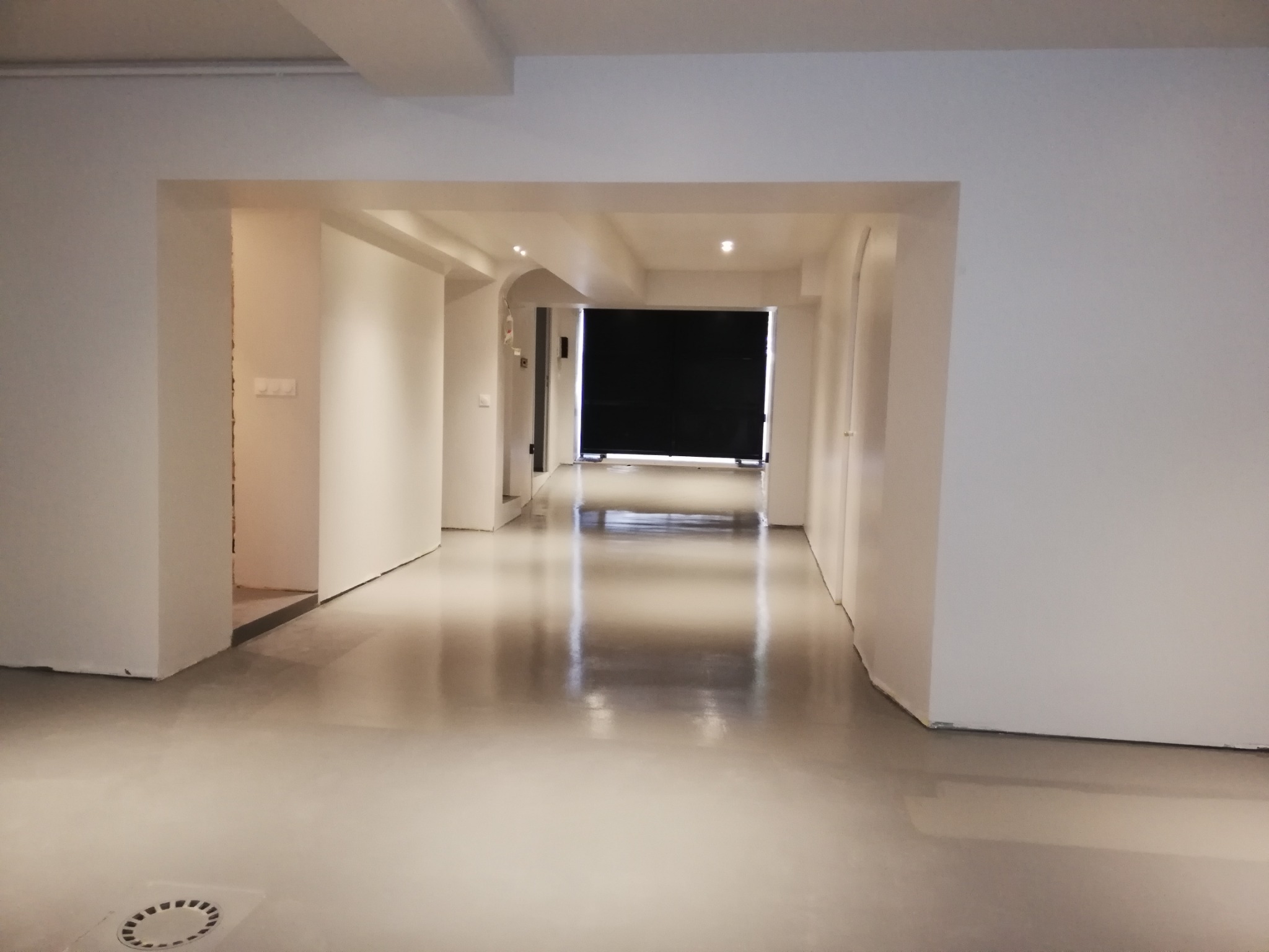 Wnętrze korytarza z gładką, jasną posadzką żywiczną, widok na otwarte przejścia między pomieszczeniami, białe ściany i sufit, z kratką ściekową w podłodze w dolnym lewym rogu.