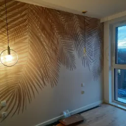 Pokój z tapetą w liście palmowe, drewnianą podłogą i dwoma nowoczesnymi lampami wiszącymi z metalowymi obręczami i widocznymi żarówkami. Przy ścianie leżą czarne listwy przypodłogowe i karton.