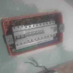 Krzysztof Baczyński - Otwarta skrzynka elektryczna z widocznymi bezpiecznikami, przewodami i elementami montażowymi w ścianie, z widocznym fragmentem instalacji wychodzącym ze ściany.