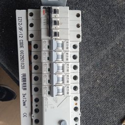 Krzysztof Baczyński - Szereg wyłączników nadprądowych Eaton Xpole Home B16/1 i B10/1 na szorstkim, ciemnym tle, obok listwa z oznaczeniem przewodu 3x12mm2, widoczne ślady użytkowania.