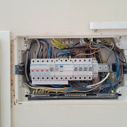 Krzysztof Baczyński - Otwarta skrzynka elektryczna w ścianie z widocznymi przewodami, bezpiecznikami i listwą zaciskową, obok znajduje się biała obudowa z żółtą naklejką 'ACZ 2 SK1', ściana w odcieniach beżu.