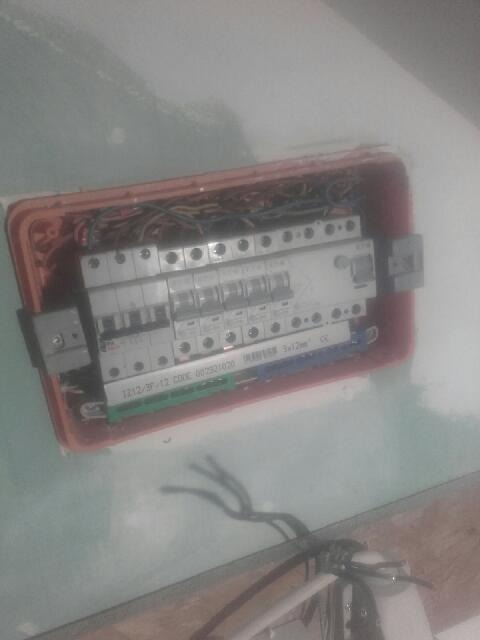 Otwarta skrzynka elektryczna z widocznymi bezpiecznikami, przewodami i elementami montażowymi w ścianie, z widocznym fragmentem instalacji wychodzącym ze ściany.