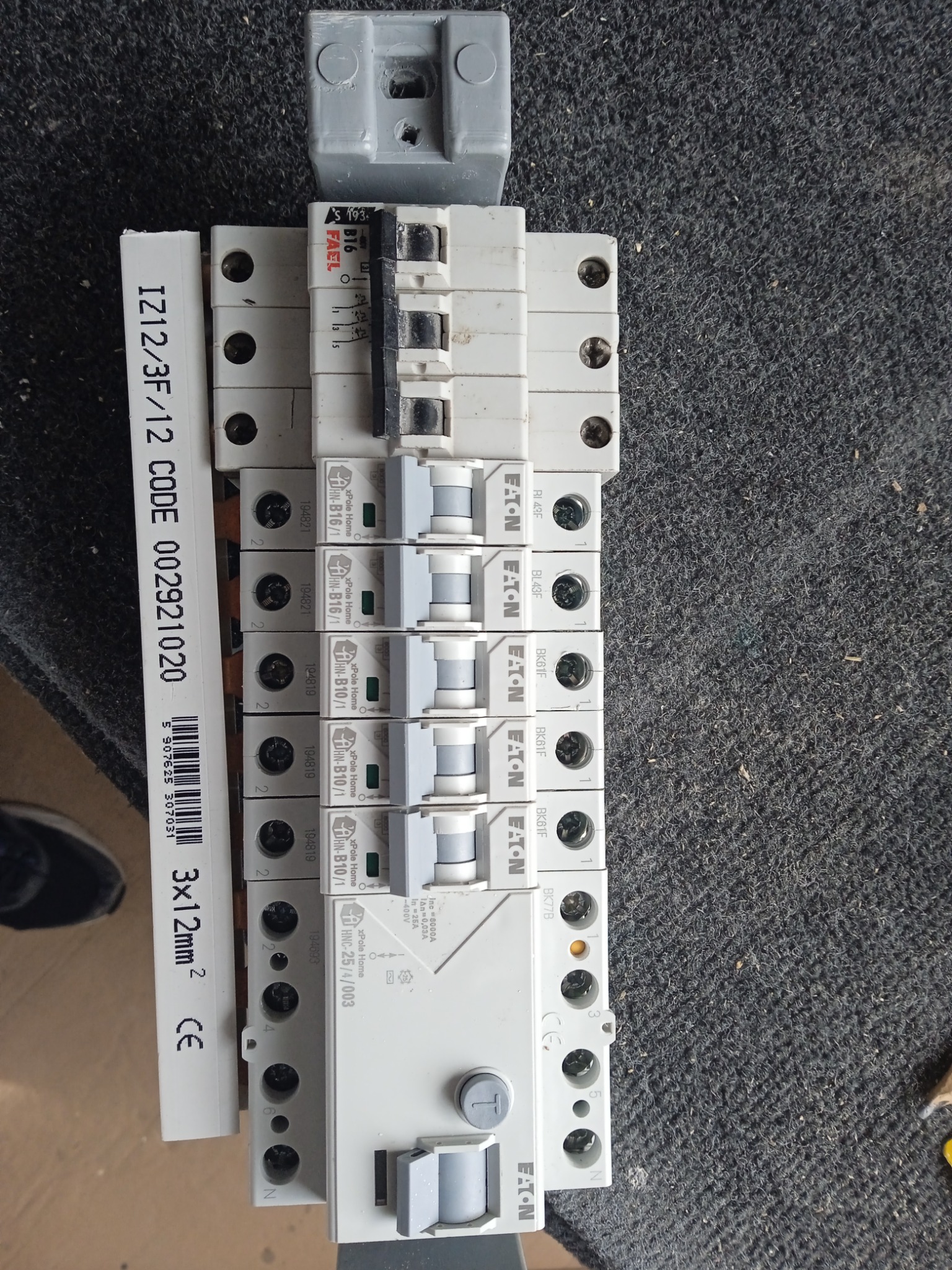 Szereg wyłączników nadprądowych Eaton Xpole Home B16/1 i B10/1 na szorstkim, ciemnym tle, obok listwa z oznaczeniem przewodu 3x12mm2, widoczne ślady użytkowania.