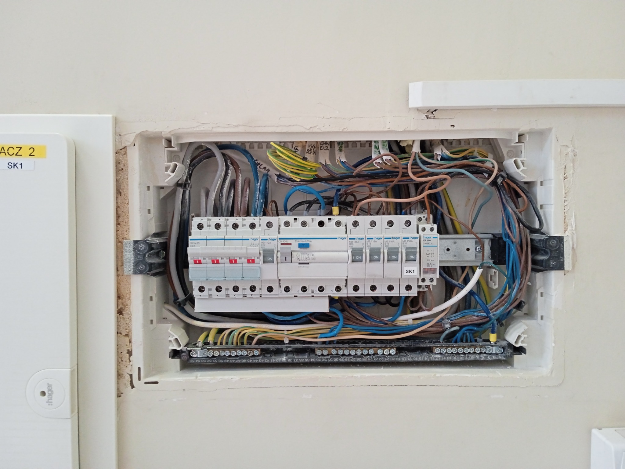 Otwarta skrzynka elektryczna w ścianie z widocznymi przewodami, bezpiecznikami i listwą zaciskową, obok znajduje się biała obudowa z żółtą naklejką 'ACZ 2 SK1', ściana w odcieniach beżu.