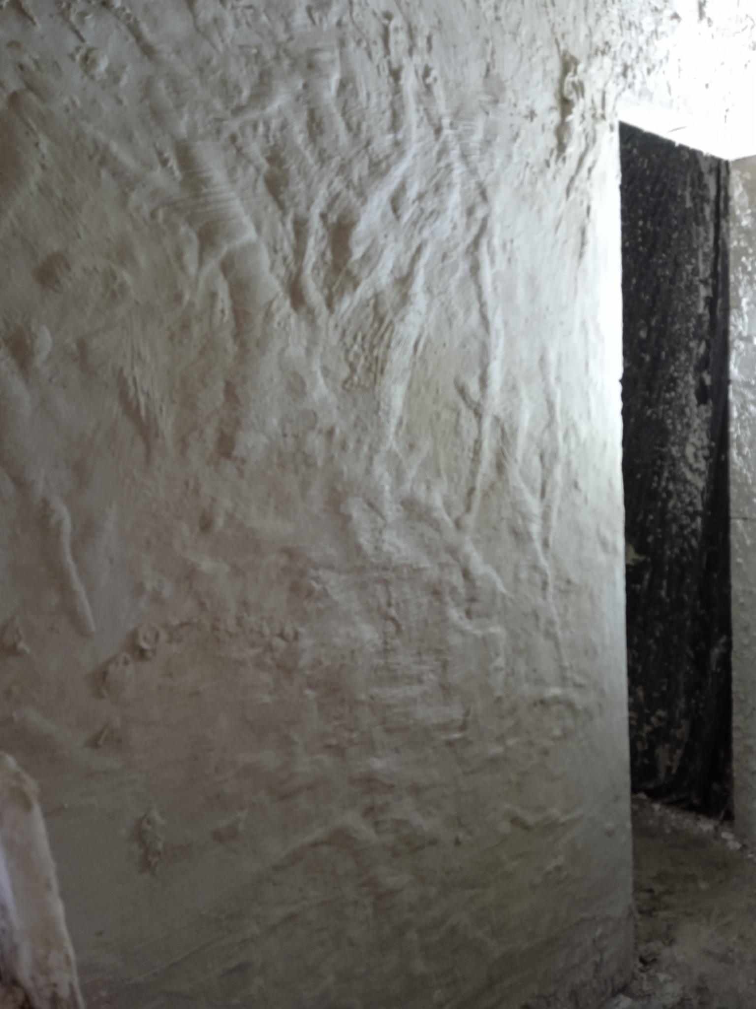 Ściana z nałożonym tynkiem w surowym stanie, widoczne nierówności i faktura, obok otwór drzwiowy z czarną folią ochronną, na podłodze resztki zaprawy.