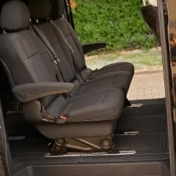 Wnętrze czarnego busa z trzema rzędami czarnych, materiałowych siedzeń, widoczne szyny mocujące fotele w podłodze i otwarte drzwi przesuwne.